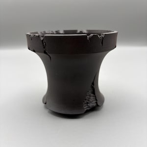 Puede incluir: Un recipiente cilíndrico acampanado de color negro mate con un borde superior ancho y abierto de bordes irregulares y dentados. La superficie exterior muestra un acabado liso que contrasta con una grieta vertical profunda.
