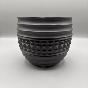 May include: A matte black cylindrical planter featuring a textured exterior with horizontal ridges along the top and bottom sections, and a central band composed of a repeating geometric pyramid pattern around the entire circumference of the vessel.