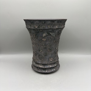 May include: A dark gray vase with a wide, flared rim and a tapered body. The surface features a heavily textured, cratered finish with metallic, bronze-toned highlights scattered throughout the uneven, porous-looking exterior pattern.