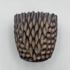 May include: A dark brown ceramic pot with a textured surface. The pot features a repeating, scale-like pattern across its surface, creating a tactile and visually interesting design. The pot has a rounded shape and a slightly flared rim.