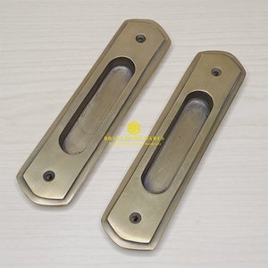 May include: Two antique brass cabinet door pulls with a rectangular shape and a long, oval-shaped cutout in the center.