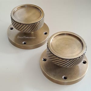 May include: Two brass-toned cabinet knobs. Each knob has a circular base with three screw holes and a rounded top. The top section features a textured, diagonal striped pattern. The knobs are likely for furniture or home decor.