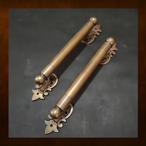 Puede incluir: Dos tiradores de puerta de latón adornados con un diseño de volutas decorativas. Los tiradores son largos y cilíndricos con un extremo redondeado.