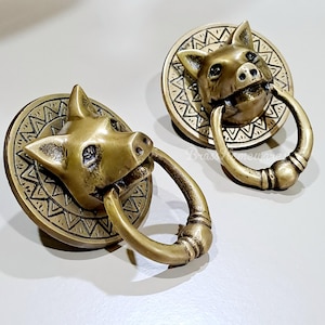 May include: Two antique brass door knockers. Each knocker features a pig head with a ring in its mouth. The pig heads are mounted on a round plate with a geometric pattern around the edge. The knockers are a warm, golden color.