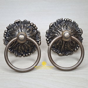 May include: Two antique brass door pulls with ornate floral designs and a ring handle. The pulls have a distressed finish and are perfect for adding a touch of vintage charm to any home.