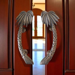 May include: Ornate bronze palm tree door handles fixed to a dark red wooden door. The handles feature detailed palm fronds and a textured trunk design. The door is ajar, offering a view of the interior space beyond.