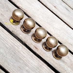 以下が含まれることがあります： 丸くて滑らかなデザインのアンティーク真鍮製のキャビネットノブが5つ。ノブは木製の表面に一列に並んでいます。