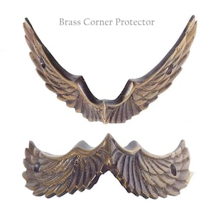 May include: Brass corner protector with a wing design. The protector is shown in two views, one from the side and one from the front.