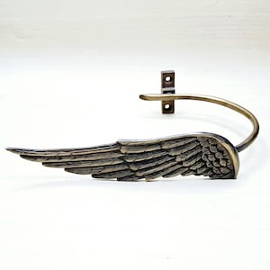May include: A bronze-colored metal wall hook shaped like a stylized wing. The wing has detailed feather textures and curves upward to meet a curved bar and a mounting plate with two holes.