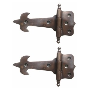May include: Two antique-style, ornate, brass hinges with a fleur-de-lis design. The hinges are closed and have a decorative, pointed finial.