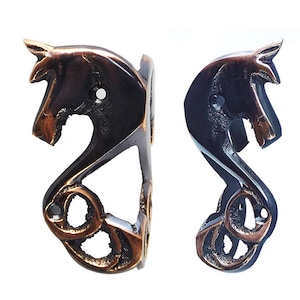May include: Two bronze-coloured, horse-head shaped decorative metal pieces. The horse heads have a stylized design with a curved neck and detailed mane. Likely cabinet hardware or decorative accents.