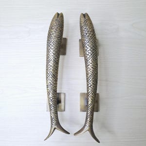 May include: A pair of bronze-toned door handles, each shaped like a fish. The handles feature intricate scale detailing and are fixed to square plates. The fish tails are oriented downwards. The background is light wood.
