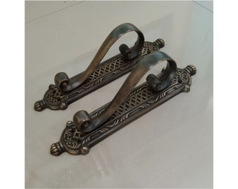 Pode incluir: Duas maçanetas de porta antigas de bronze. Cada maçaneta apresenta uma pega arqueada e curva sobre uma base retangular com um design vazado ornamentado. As bases têm peças finais decorativas com volutas e motivos florais.