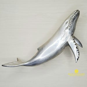 May include: A silver whale sculpture with a detailed, textured surface. The whale is positioned vertically with its tail fin raised.