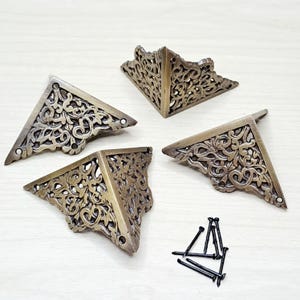 May include: Four antique bronze corner brackets with intricate floral patterns. Each bracket is triangular and has pre-drilled holes for fixing. A small collection of black nails is also visible, suitable for furniture restoration or decorative use.