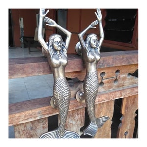 Könnte beinhalten: Zwei antike Messing-Türgriffe in Form einer Meerjungfrau mit erhabenen Details. Die Meerjungfrauen sind mit erhobenen Armen über ihren Köpfen und ihren Schwänzen, die sich hinter ihnen zusammenrollen, dargestellt.