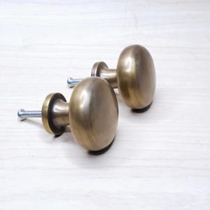 Op de afbeelding: Twee antieke messing kastknoppen met ovale handgrepen en schroefbouten. De knoppen hebben een warme, metallic afwerking en zijn geplaatst op een lichtgekleurd houten oppervlak. Deze knoppen zijn geschikt voor meubelrestauratie.