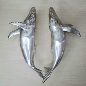 Op de afbeelding: Twee gepolijste zilverkleurige deurklinken in de vorm van een walvis. Elke handgreep heeft een gedetailleerd ontwerp van een walvis, inclusief vinnen en een getextureerde kop. De handgrepen zijn ontworpen om verticaal te worden gemonteerd.