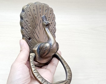 Könnte beinhalten: Bronzener Türklopfer in Form eines Pfaus mit detailliertem Federdesign. Der Türklopfer zeigt einen Pfau mit einem gebogenen Griff. Der Klopfer ist auf einer rechteckigen Rückplatte mit fächerförmigem Design montiert.