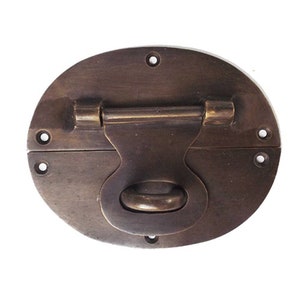 May include: Oval antique bronze trunk latch with a hinged handle and a circular pull. The latch is secured with a hinged bar and is mounted with six small screws. The metal has a weathered appearance.
