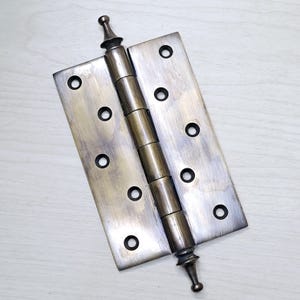 May include: A bronze-toned door hinge with a decorative finial at each end. The hinge has a series of cylindrical links connecting two rectangular plates, each with multiple circular screw holes. The metal has a slightly aged appearance.