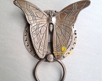 Puede incluir: Un llamador de puerta de latón con forma de mariposa con detalles intrincados. La mariposa tiene una base redonda y decorativa con un anillo para llamar.