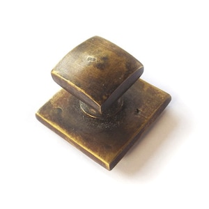 May include: A square brass cabinet knob with a slightly raised, square top. The knob has a worn, aged finish.