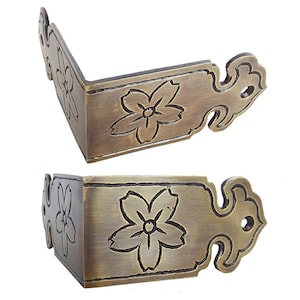 Puede incluir: Dos soportes de esquina de latón antiguo con un diseño floral. Cada soporte tiene un diseño decorativo de volutas y un patrón floral en relieve.