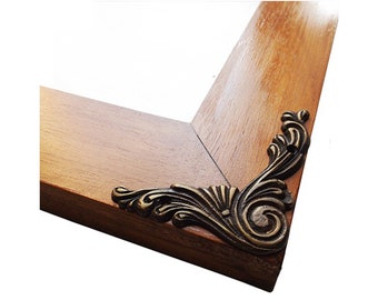 May include: A wooden picture frame with ornate metal corner accents. The frame is a light brown color and the accents are a gold color.
