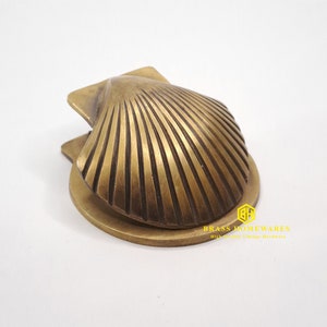 Może przedstawiać: Miedziany klamka do drzwi w kształcie muszli św. Jakuba. Klamka ma szczegółowy, fakturowany wzór i jest zamontowana na okrągłej podstawie.