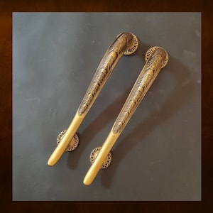 May include: Two ornate gold door handles with intricate designs. The handles are long and slender with a rounded end. They are mounted on a dark grey surface.