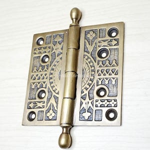 May include: An antique brass door hinge with an ornate, engraved design. The hinge features a rectangular plate with a detailed pattern of floral and geometric shapes, and a cylindrical barrel with a decorative finial.