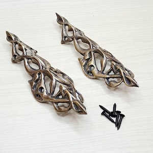 May include: Two bronze-coloured, ornate cabinet pulls with an openwork design. The pulls have a detailed, organic pattern. A small pile of black screws is included, suggesting installation hardware. The pulls are on a light-coloured surface.