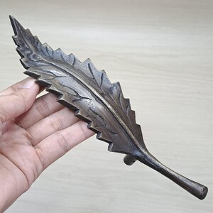 Op de afbeelding: Een bronzen, bladvormig decoratief object. Het blad heeft gedetailleerde nerven en een getextureerd oppervlak. De steel van het blad is licht gebogen en de randen zijn gekarteld. Het object wordt in een hand gehouden.