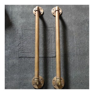 Puede incluir: Dos tiradores de puerta de latón con diseño de tubería. Los tiradores están montados en una pared gris oscura.