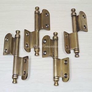 Puede incluir: Cuatro bisagras antiguas de latón con remates decorativos. Cada bisagra tiene forma rectangular con bordes redondeados y múltiples orificios para tornillos. Las bisagras son de color latón cálido y envejecido.