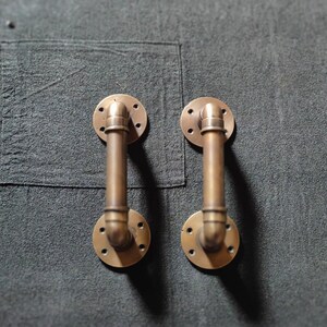 Puede incluir: Dos tiradores de puerta de latón antiguo con un diseño de tubería. Los tiradores están montados en una superficie gris oscuro.