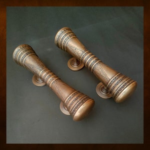 Puede incluir: Dos tiradores de puerta de cobre adornados con diseños intrincados. Los tiradores tienen forma de cilindros con un extremo redondeado y una base plana.