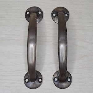 Könnte beinhalten: Zwei antike Türgriffe aus Bronze. Jeder Griff hat eine gebogene Stange und zwei kreisförmige Montageplatten mit Schraubenlöchern. Die Bronze hat ein verwittertes Aussehen mit Farb- und Texturvariationen.