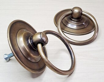May include: Two antique brass-toned drawer pulls with ring handles. Each pull features a circular backplate and a rounded ring handle. One pull has a visible screw. The hardware is designed for furniture restoration or decorative use.