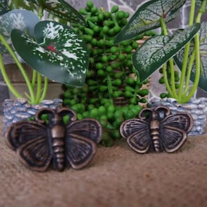 May include: Two antique bronze butterfly drawer pulls with intricate details. The pulls are set against a backdrop of green plants.