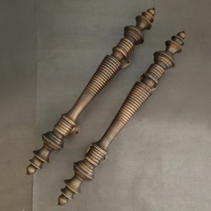 Puede incluir: Un par de tiradores de puerta de latón antiguo con diseños intrincados y ornamentados. Los tiradores son largos y delgados, con una punta puntiaguda en la parte superior y un pomo decorativo en la parte inferior.