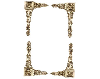 May include: Set of four gold-tone metal corner brackets with ornate designs. Each bracket has a square center with a floral design and a decorative scrollwork pattern.