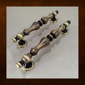 Könnte beinhalten: Zwei verzierte Türgriffe in Bronzefarbe mit schwarzen Akzenten. Die Griffe weisen ein detailliertes, Vintage-Design mit geschwungener Form und dekorativen Elementen auf. Die Beschläge sind wahrscheinlich für einen Schrank oder eine Tür.