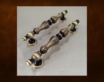 Pode incluir: Duas puxadores de porta ornamentados em tom bronze com detalhes em preto. As puxadores apresentam um design vintage detalhado com uma forma curva e elementos decorativos. A ferragem é provavelmente para um armário ou porta.