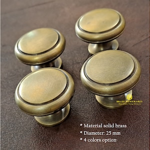 以下が含まれることがあります： 4つのアンティークブラスキャビネットノブ。隆起した縁があり、直径25mmです。ノブは茶色の革の表面にあります。 *素材：無垢の真鍮 *直径：25mm *4色からお選びいただけます