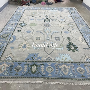 May include: Large rectangular area rug with a light gray field and a blue border. The rug features a detailed floral and geometric pattern in shades of blue, green, and cream. The text "Knot & ufti" is visible on the rug.