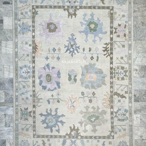 May include: A rectangular, beige-colored rug with a floral pattern. The rug features a central design of large, stylized flowers in shades of blue, purple, and green, framed by a border with geometric and floral motifs. The text "KnotAndTuft" is visible.