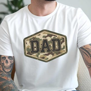 May include: White t-shirt featuring a camouflage design with the word "DAD" in bold, dark letters. The text is inside a hexagonal frame with a distressed look. The shirt is a casual wear item.