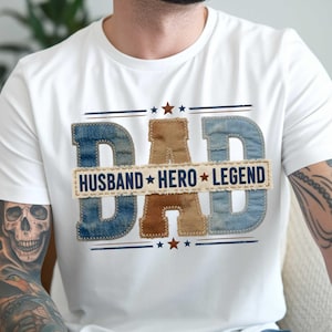 May include: White t-shirt with the word "DAD" in denim and brown letters. The word "DAD" is above a rectangle with the words "HUSBAND HERO LEGEND". The design includes blue and brown stars and lines.